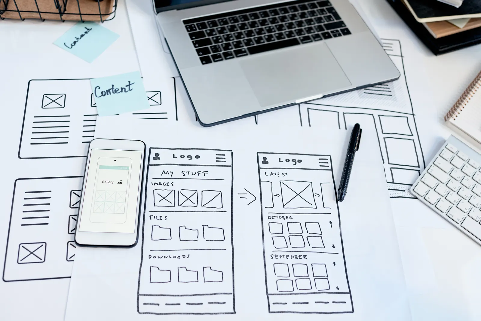 Hand-drawn app screen sketches on paper beside a phone and a laptop